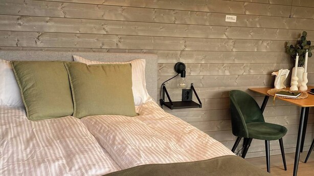 Un lit bien fait, recouvert d’une couvre-lit à rayures et agrémenté de coussins verts, une petite table avec une bougie et une plante, et une chaise avec un coussin foncé.