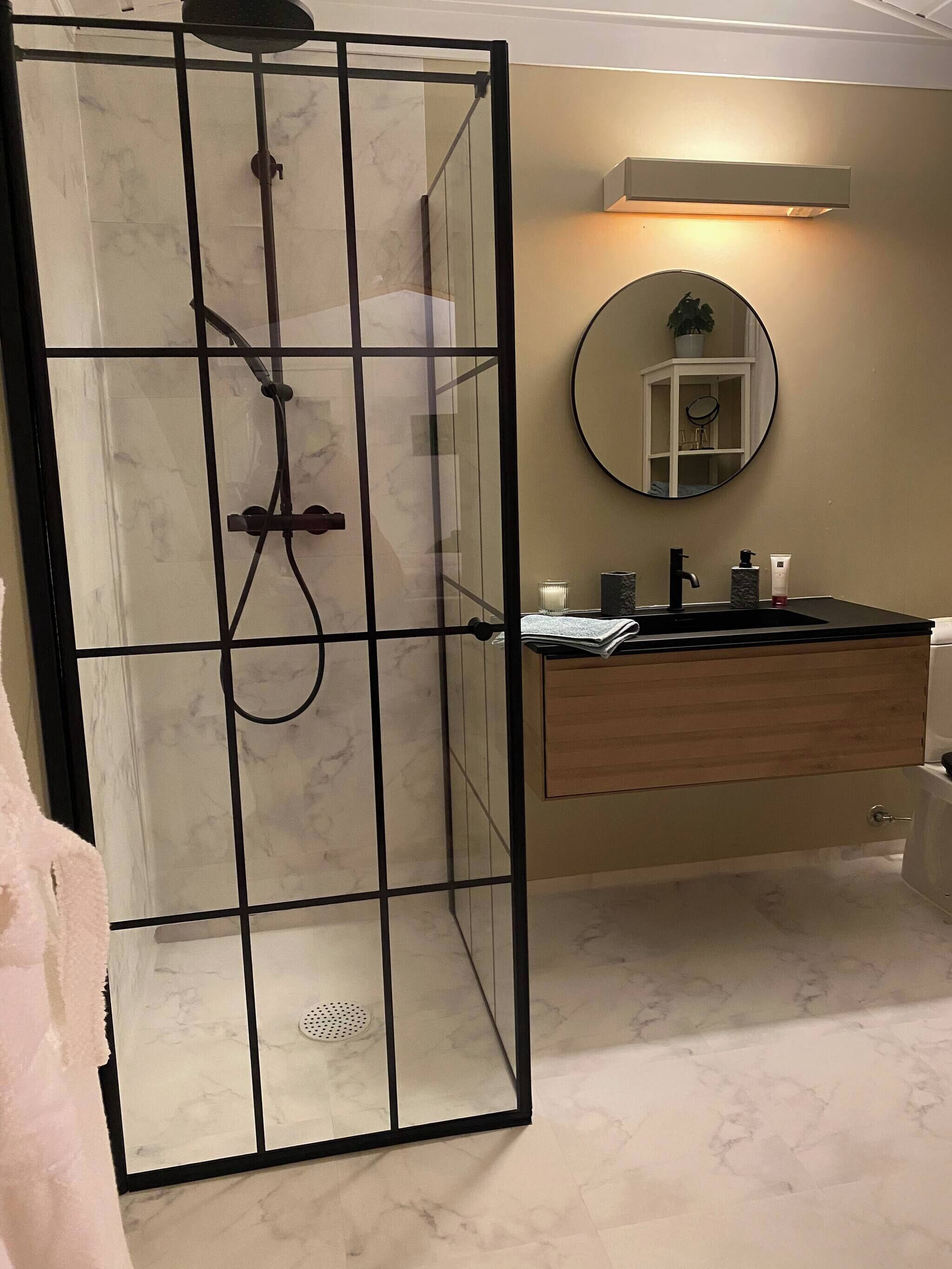 A modern bathroom with a glass-enclosed shower, a wooden vanity, a round mirror, and a marble-tiled floor.