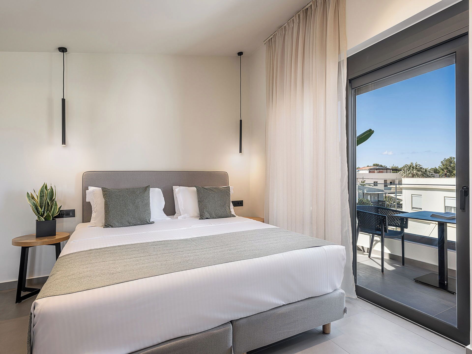 A modern bedroom with a large bed, a small side table with a plant, and a view of the outside balcony.