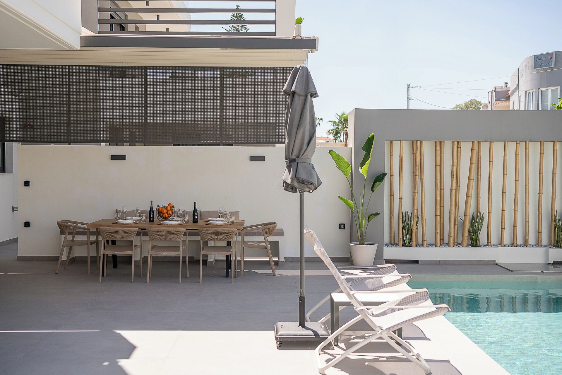 Ein moderner Essbereich im Freien mit Pool, der einen Tisch für vier Personen, eine überdachte Lounge-Ecke und eine Bambuswand zeigt.