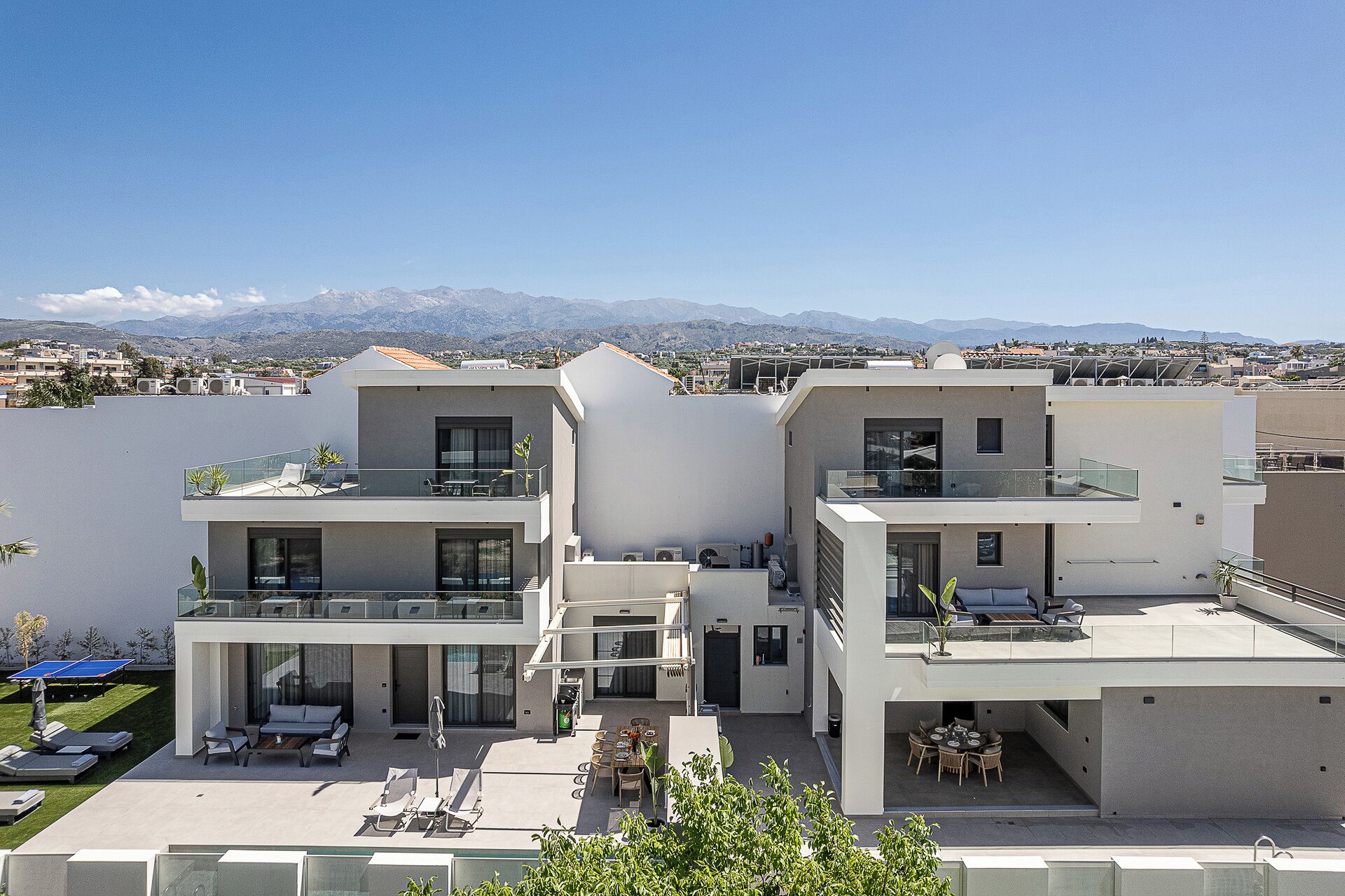 A modern two-story residential building with a rooftop terrace, large windows, and a view of distant mountains.