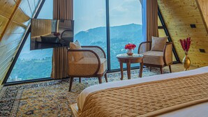 A bedroom with a large window, a bed, a chair, a small table, and a ceiling fan. - Alaya Stays Echo By The Lakes (Bhimtal)