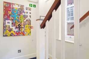 A hallway with a colorful abstract painting on the wall, a staircase with a wooden handrail, and a window with a view of a brick building. - Cozy Studio in the Heart of the City of London A (London)