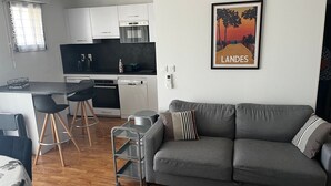 A modern kitchen with white cabinets, a black countertop, and a dining area with a gray sofa and black chairs. - Apartment 'Les Balcons du Soleil' – Ideal for Spa Guests - Conciergerieland (Saint-Paul-lès-Dax)