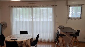 A dining area with a round table, chairs, a cabinet, and a window with curtains. - Apartment 'Les Balcons du Soleil' – Ideal for Spa Guests - Conciergerieland (Saint-Paul-lès-Dax)