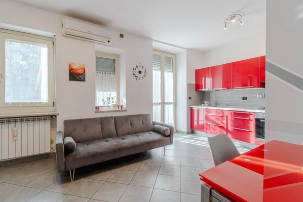 A modern living room with a grey sofa, a red dining table, and a kitchen with red cabinets.