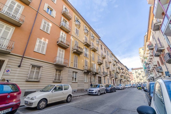 A street lined with multi-story buildings, parked cars, and a clear sky.