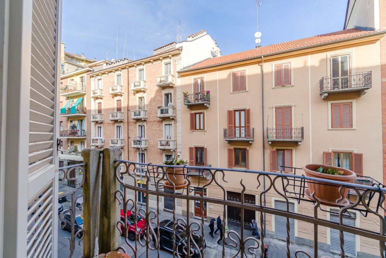 Ein Balkon mit schmiedeeisernen Geländern, Terrakottatöpfen und Blick auf mehrstöckige Gebäude mit Balkonen und Fensterläden.