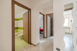 A hallway with a bathroom on the left, a bedroom with a red bed in the middle, and a white wall with a unique design on the right.