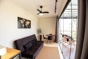 Una sala de estar moderna con un sofá oscuro, una mesita, un ventilador de techo y un amplio ventanal con vistas al exterior.