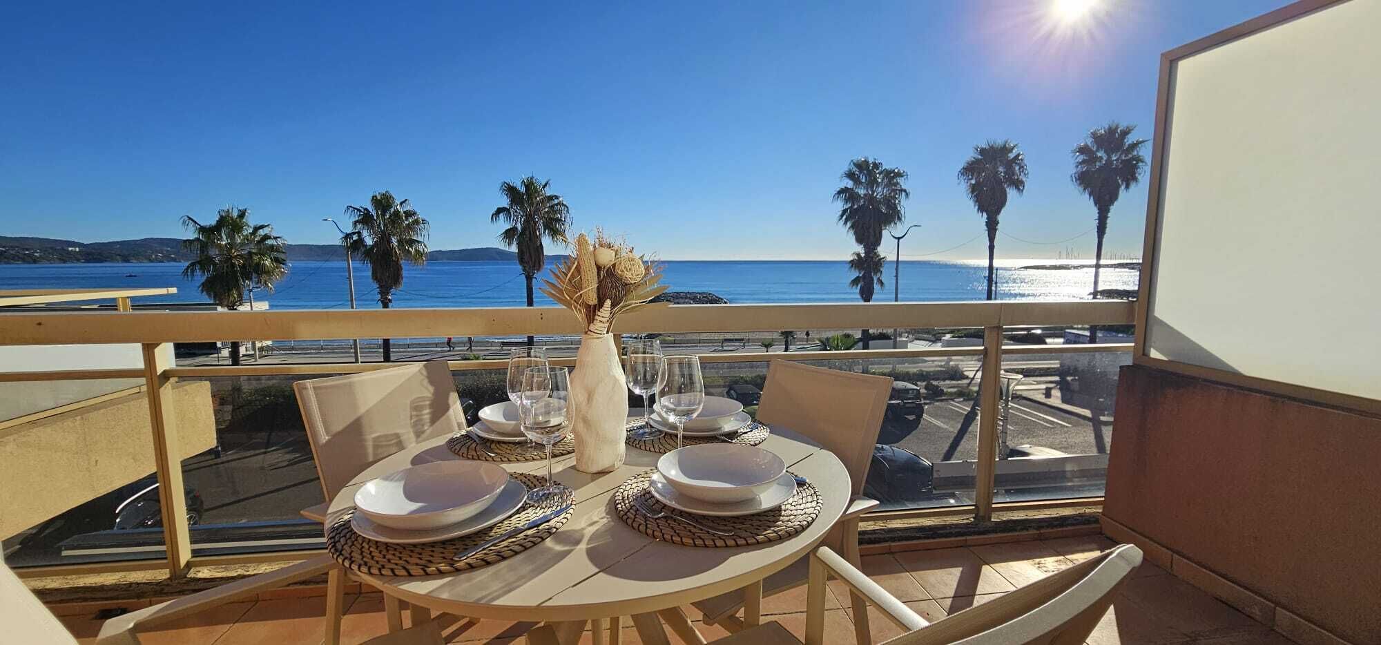 Une table à manger dressée pour quatre personnes, avec des assiettes, des verres et un vase, sur un balcon donnant sur la mer.