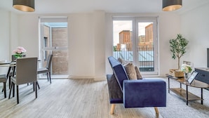 A modern living room with a blue sofa, dining table, chairs, and a view of buildings through large windows. - City View Apartment Near Manchester City Centre (Salford)