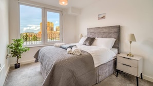 A bedroom with a large window, a bed with a grey headboard, two bedside lamps, and a small potted plant. - City View Apartment Near Manchester City Centre (Salford)