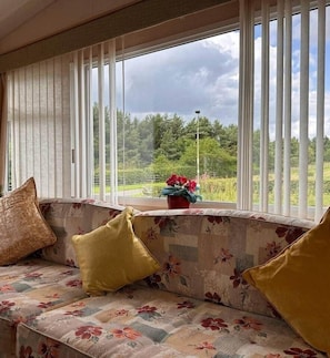 A room with a sofa facing a window with vertical blinds, a potted plant on the windowsill, and a view of a green landscape. - 4-Bedroom Cosalt Static Caravan Uddingston Glasgow (Uddingston)
