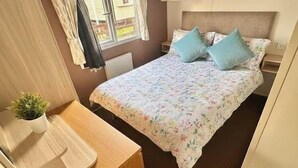 A compact bedroom with a bed, a small desk, and a window with curtains. - Delta, Bromley 4-Bedroom Caravan (Uddingston)