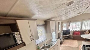 A compact kitchen with a microwave, sink, and coffee maker. There is a small dining area with a table and chairs. - Delta, Bromley 4-Bedroom Caravan (Uddingston)
