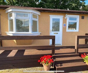 A small, single-story building with a brown exterior, a white door, and a white window. There are wooden benches in the foreground and a potted plant with red flowers. - Semi-detached chalet - Uddingston, Glasgow (Uddingston)