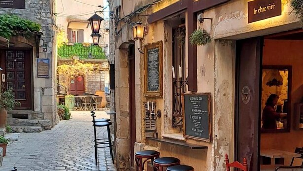 Una calle adoquinada con mesas al aire libre, edificios de piedra y un café con un cartel que dice 'le chat vire'.