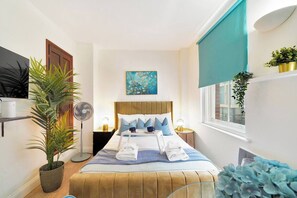 Quarto de hotel com cama, mesa de cabeceira, abajur, ventilador, janela com vista e uma planta.