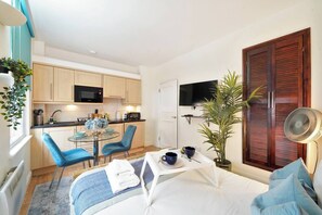 Quarto de hotel compacto com cama, uma mesinha com um ventilador, televisão e uma kitchenette com micro-ondas e armários.