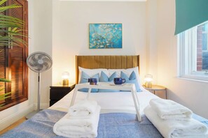 Ein ordentlich bezogenes Bett mit Kissen, ein Nachttisch mit Lampe, ein an der Wand hängendes Gemälde und ein Fenster mit Blick.