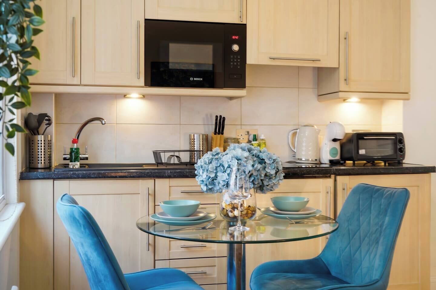 Una cocina moderna con un microondas Bosch, una mesa de vidrio con sillas azules y un jarrón con flores azules.