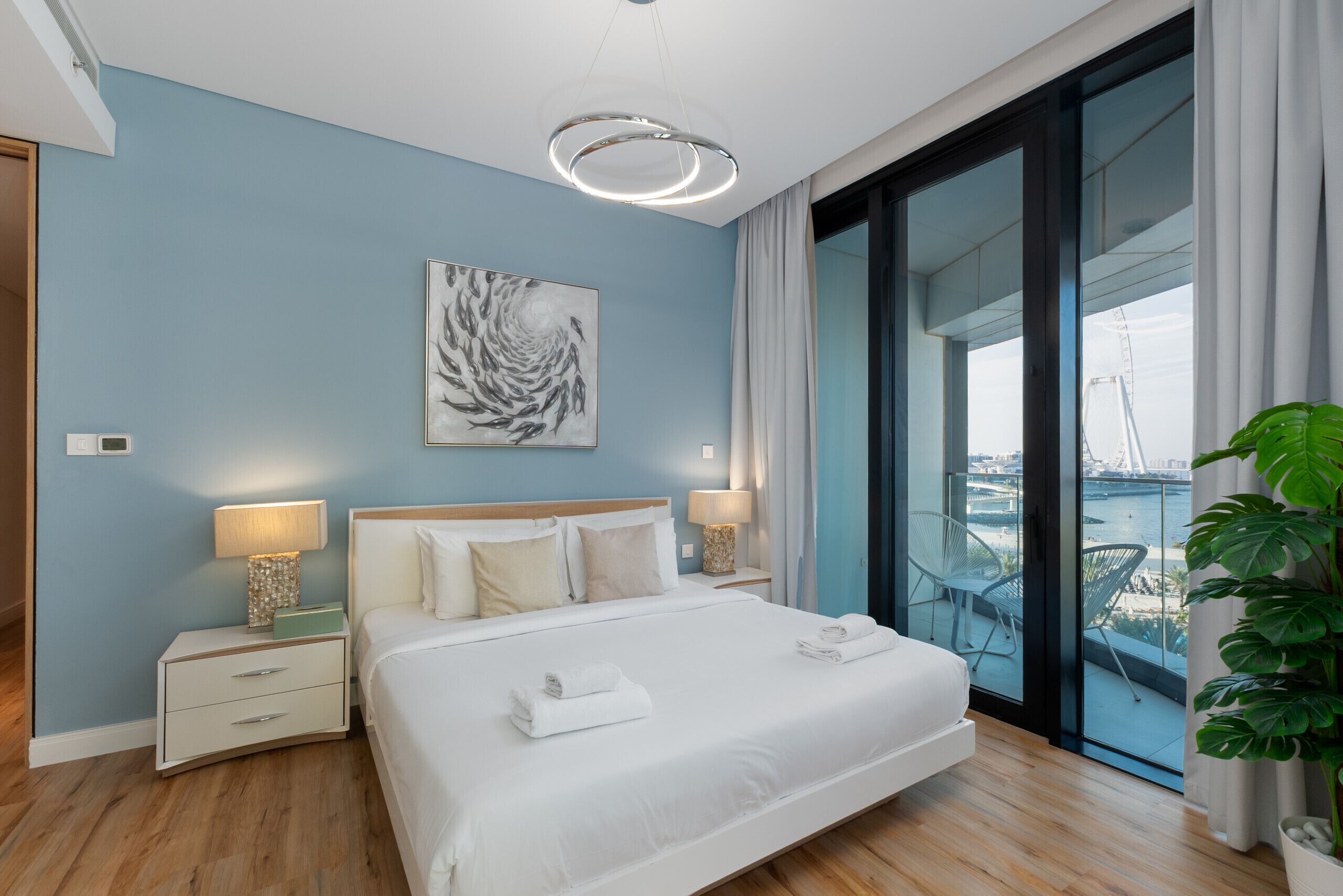 Une chambre d’hôtel moderne avec un grand lit, deux tables de chevet avec des lampes, une vue sur le paysage urbain et un balcon avec des chaises.