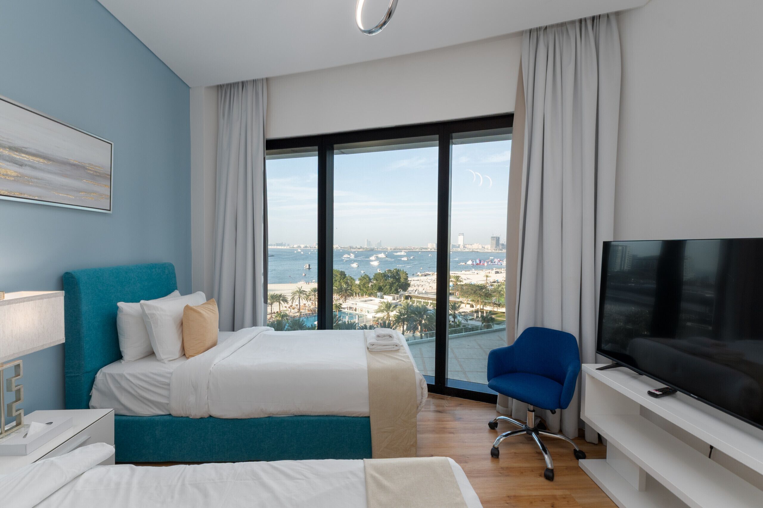 Ett hotellrum med en stor säng, en TV, en stol och utsikt över stranden och staden.