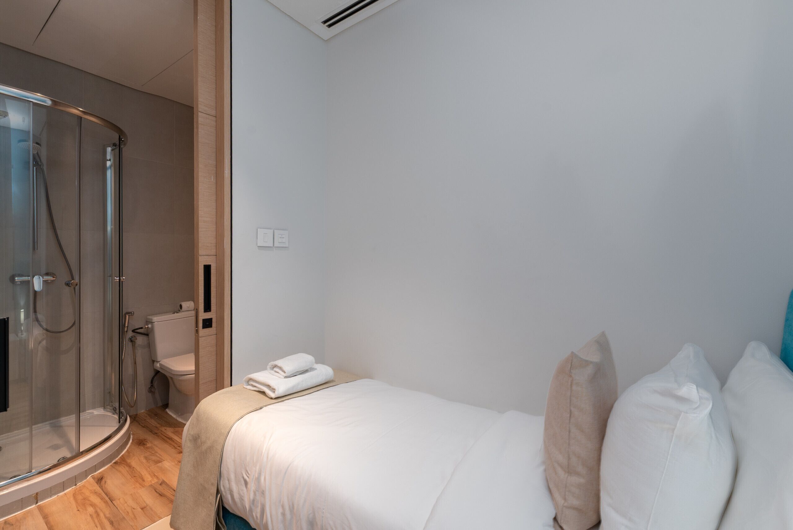 Une chambre d’hôtel moderne dotée d’une douche en verre, d’un lit avec du linge de maison blanc et d’un plancher en bois.