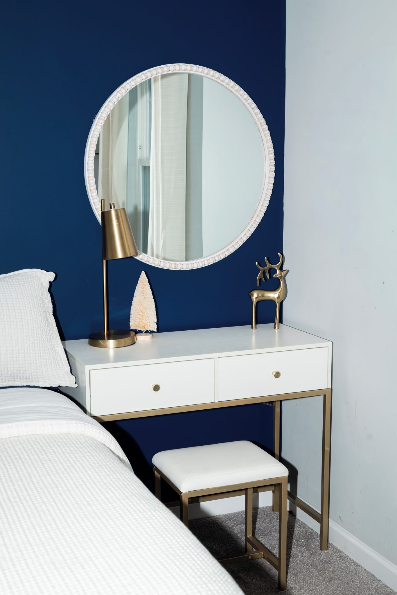 Une coiffeuse blanche avec un miroir rond, une lampe dorée et une petite figurine de cerf.