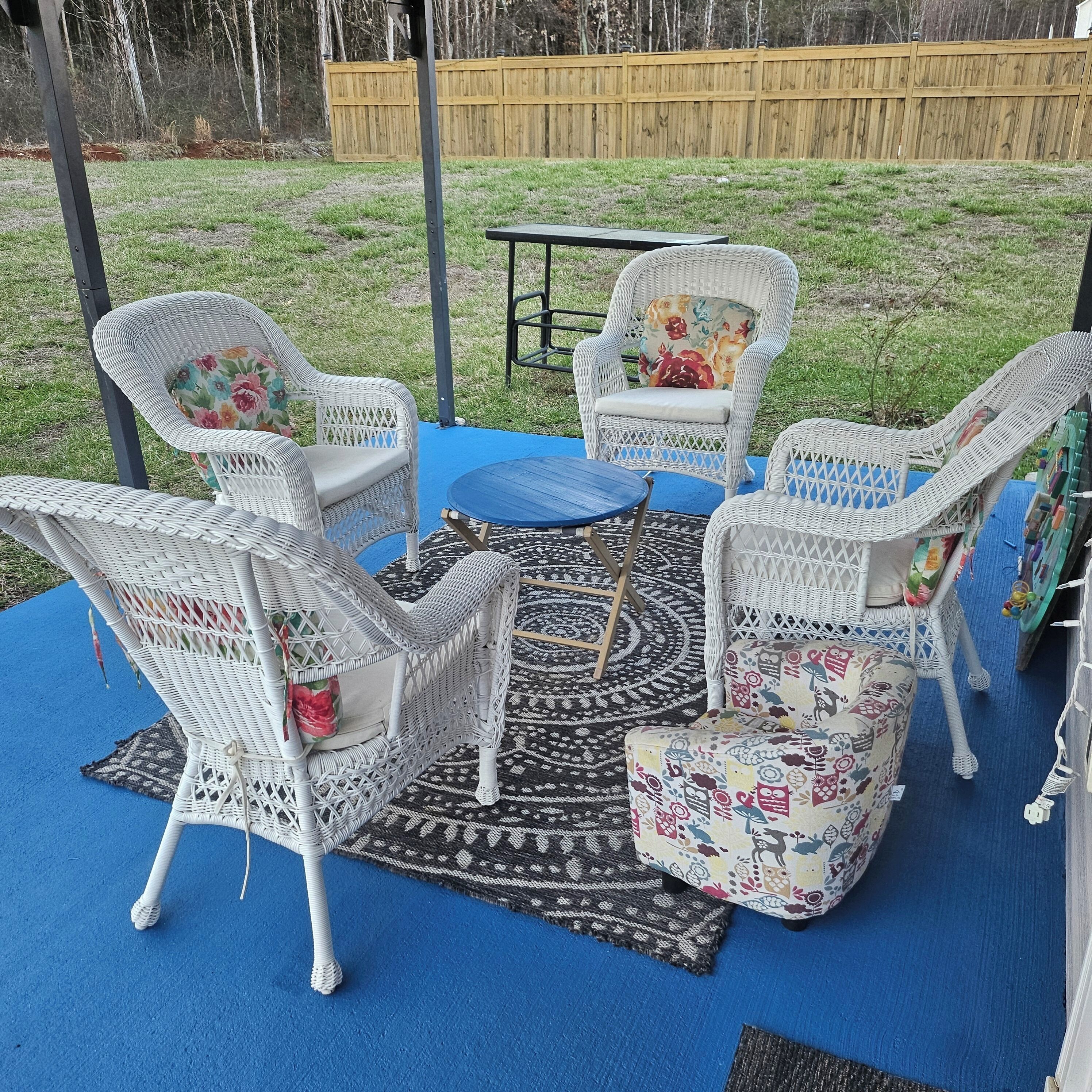 A patio area with white wicker furniture, including chairs and a table, on a blue carpet.