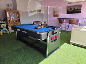 A pool table with a blue felt surface, a foosball table, a sofa, and a television mounted on the wall.