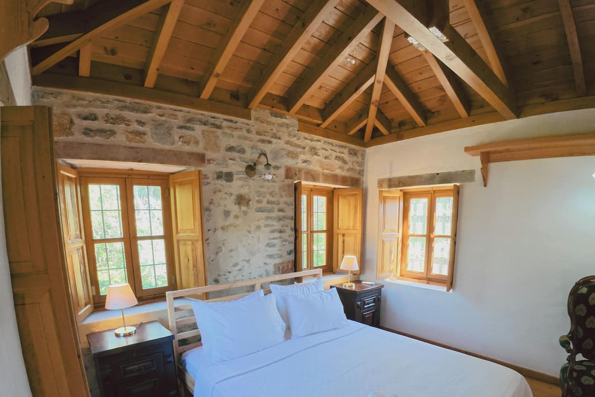 Ein Schlafzimmer mit einem großen Bett, Holzbalken, Steinwänden und zwei Fenstern.
