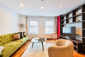 Un salon avec un canapé vert, une table basse ronde, un fauteuil beige, un téléviseur posé sur un meuble en bois et une fenêtre avec des rideaux.