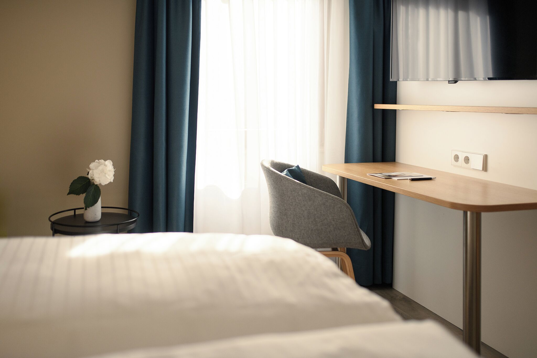 A hotel room with a bed, a desk with a chair, a small table with a vase of flowers, and a window with curtains.