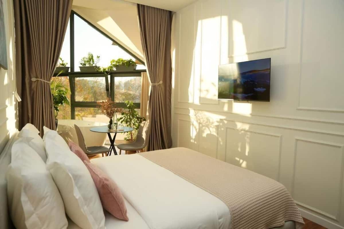 Ein Schlafzimmer mit einem Bett, einem Fernseher, einem Fenster mit Vorhängen und Blick ins Freie.