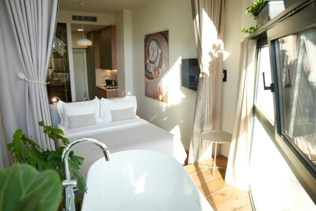 Ein modernes Hotelzimmer mit einem großen Bett, einer weißen Badewanne und Blick auf die Stadt durch das Fenster.