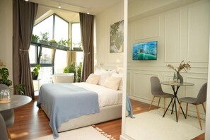 A bedroom with a bed, a dining table with chairs, a TV, and a window with curtains.
