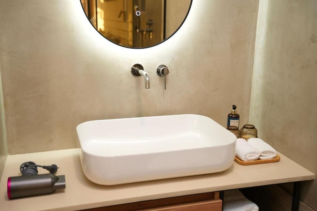 Un baño con un lavamanos rectangular blanco, un espejo redondo y un perchero con toallas dobladas.