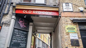 Un estrecho callejón con un pub llamado 'Old Red Cow' a la izquierda, un cartel que indica 'East Passage EC1' arriba y un anuncio de 'Deep Tissue Massage' a la derecha.