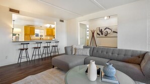 A modern living room with a grey sofa, a round glass coffee table, and a kitchen area with wooden cabinets and bar stools. - Pool / 4 BR 2 BA / Davis-Monthan AFB / Renovated (Tucson)