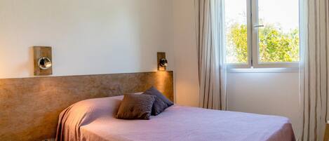 A bedroom with a bed, two wall-mounted lights, a window with curtains, and a wooden stool.