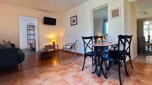 Una sala de estar con sofá, una mesa de centro y un comedor con sillas y una mesa. Hay un televisor montado en la pared y una lámpara sobre una mesita auxiliar.