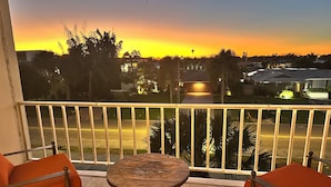 A balcony with a table and chairs, a railing, and a view of a sunset over a residential area.