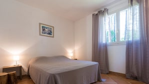A bedroom with a bed, two bedside lamps, a framed picture on the wall, and a window with curtains. - Villa du Domaine 3bed park view (Sari-Solenzara)
