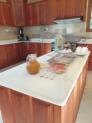 Una cocina con armarios de madera, encimera de mármol, cafetera, hervidor, jarra de agua grande y una bandeja con comida.