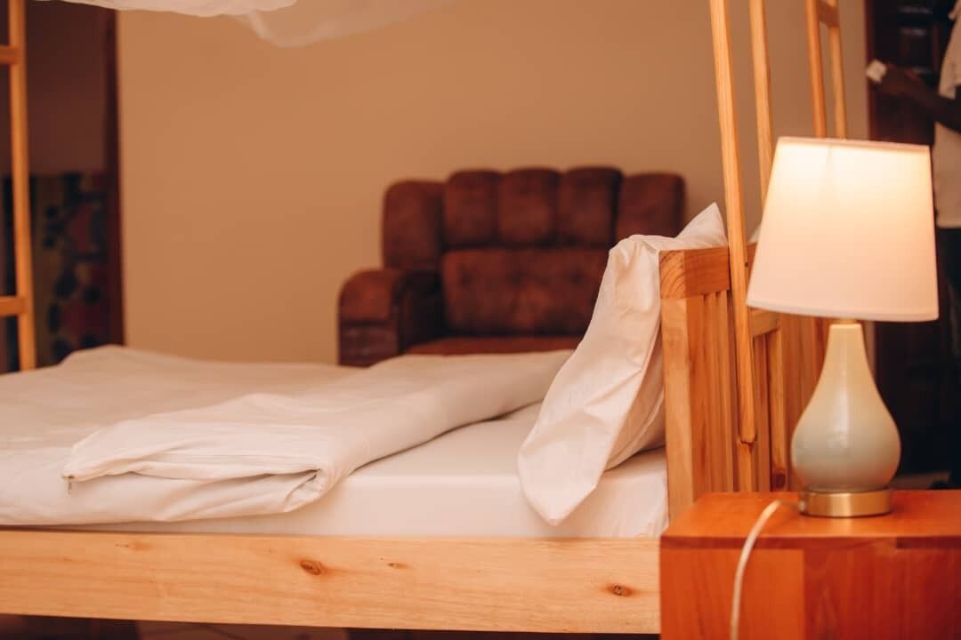 Ein hölzernes Bett mit weißen Bettwäsche, einer Nachttischlampe und einem braunen, gepolsterten Kopfteil.