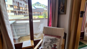 A room with a bed, a small table, a window with curtains, and a view of buildings outside.
