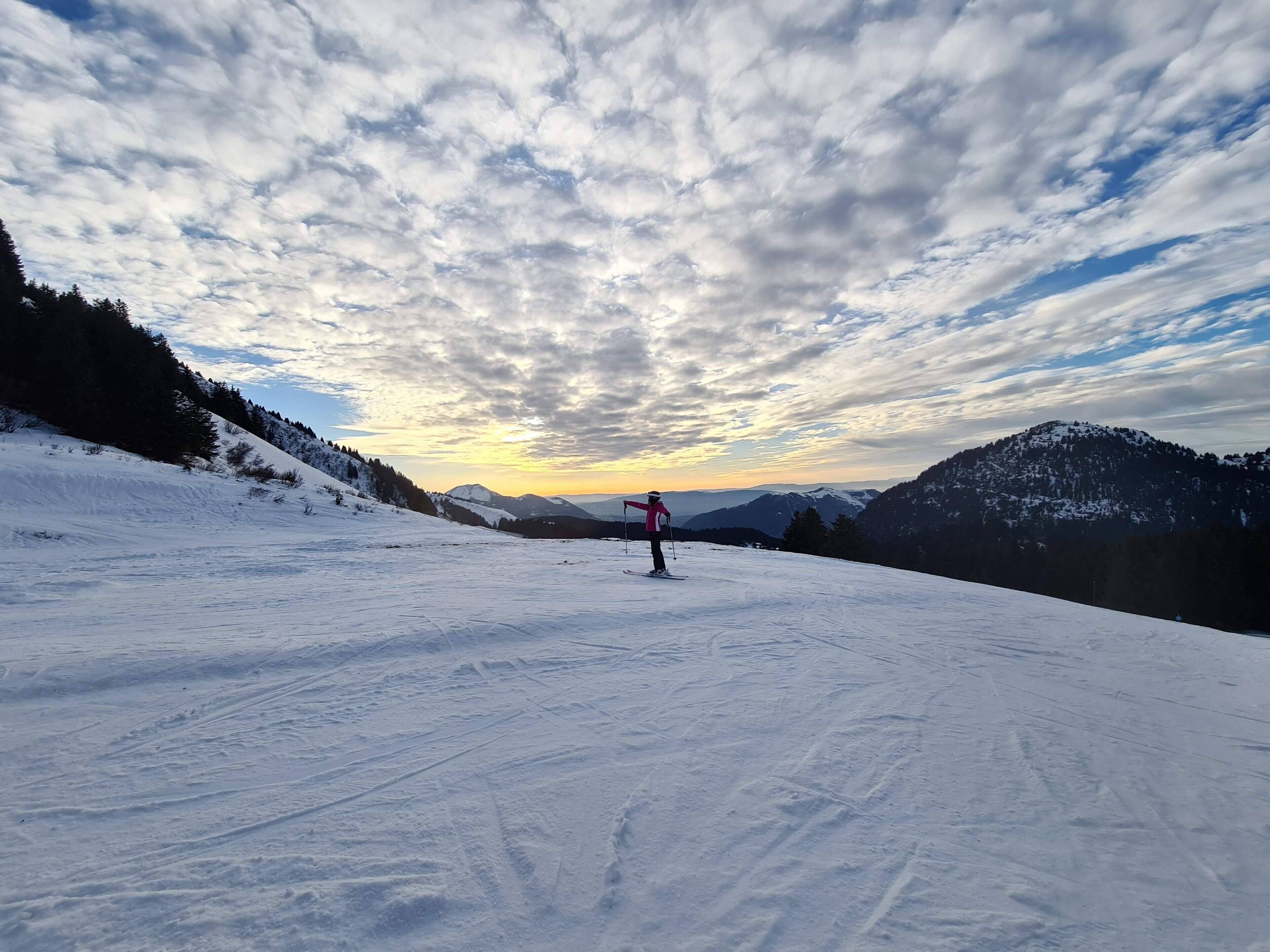 A snowy ski slope with a skier in the distance, surrounded by mountains and a partly cloudy sky.