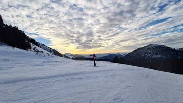 A snowy ski slope with a skier in the distance, surrounded by mountains and a partly cloudy sky.